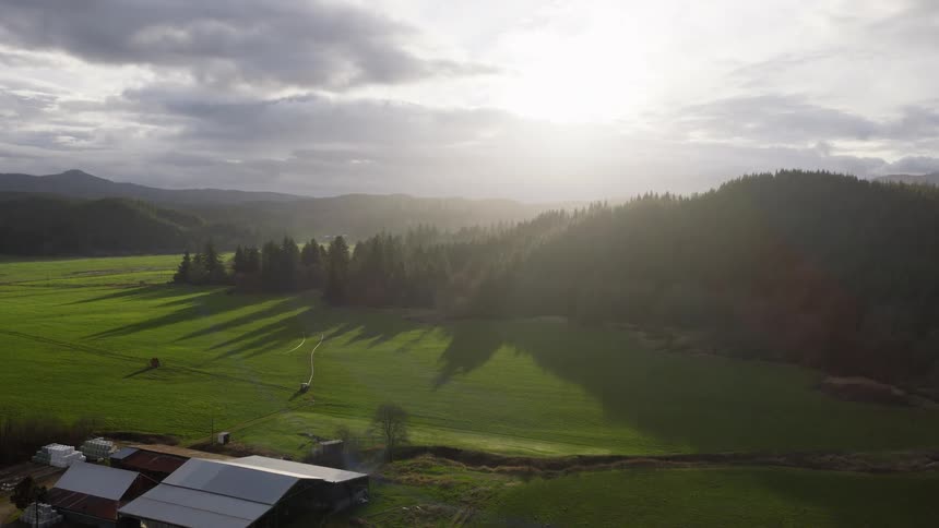 An ascending aerial view captures a beautiful farm and expansive green valley as sunlight shines through dramatic clouds over rolling hills