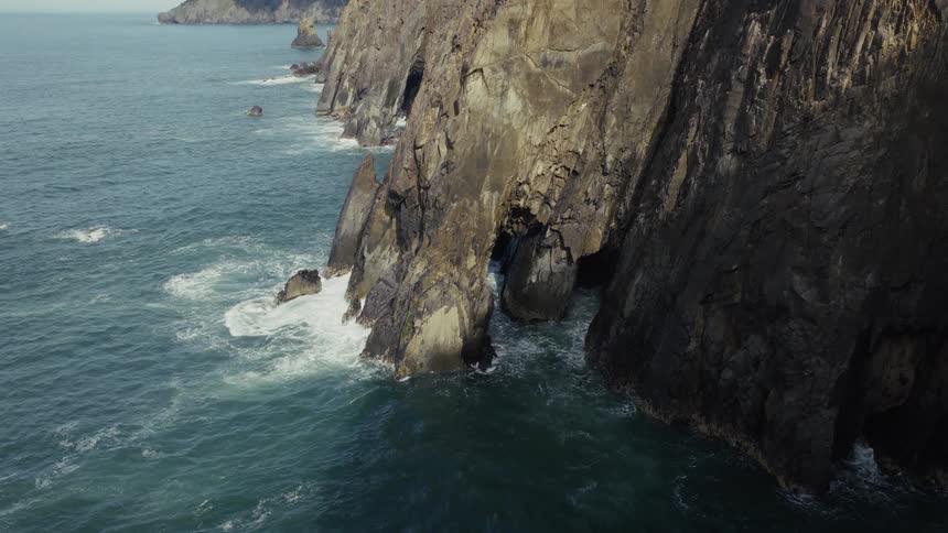 A slow reveal of a dramatic, rocky coastline shows sea caves and arches carved by the powerful ocean waves