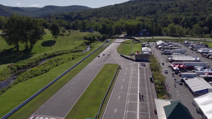 An aerial drone shot flies over the main straight of Lime Rock Park racetrack during a sunny day event, with classic race cars on the track and spectators in the paddock