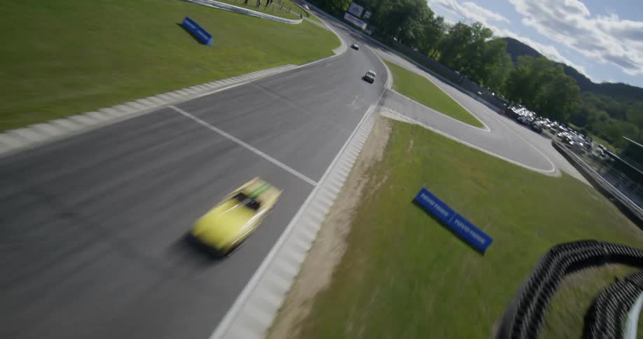 A high-speed FPV drone dynamically follows a group of vintage sports cars racing around a scenic, winding track