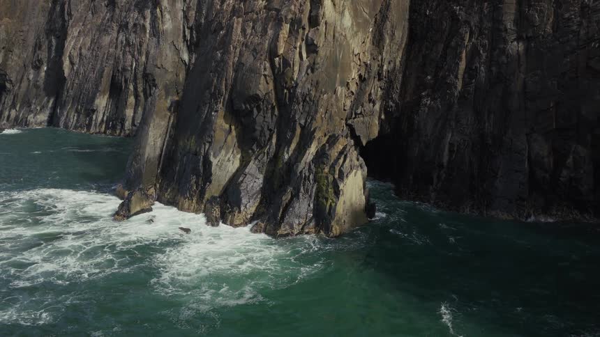 A drone pulls back from a dramatic sea arch to reveal a massive rocky cliff face pounded by ocean waves on a sunny day