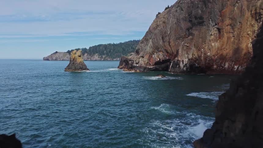 An aerial view flies alongside a dramatic, rocky coastline, passing through towering sea stacks in the ocean on a beautiful day