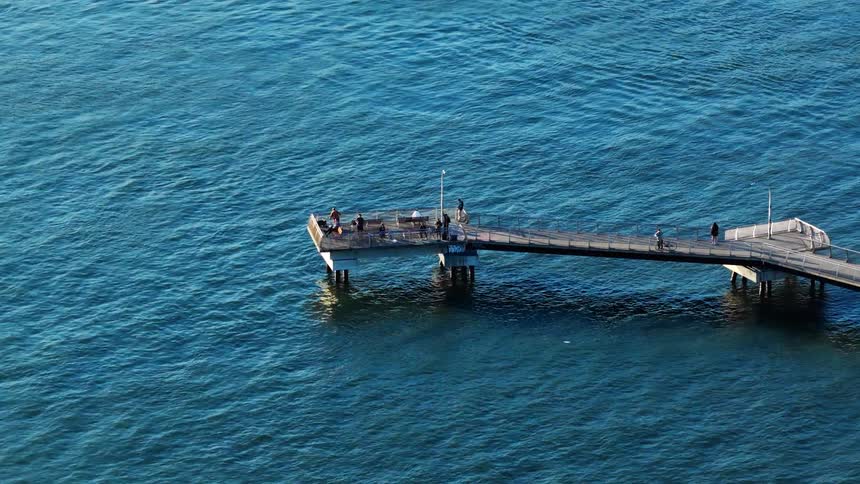 A serene high-angle shot captures people relaxing and fishing from a pier extending into the vast, calm blue ocean