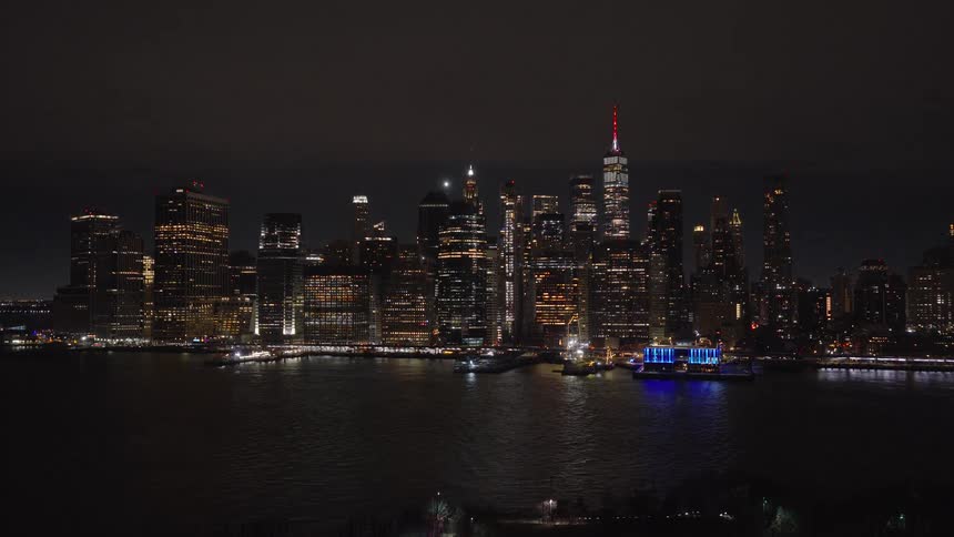 Aerial drone slowly pushes forward over the East River towards the illuminated Lower Manhattan skyline at night