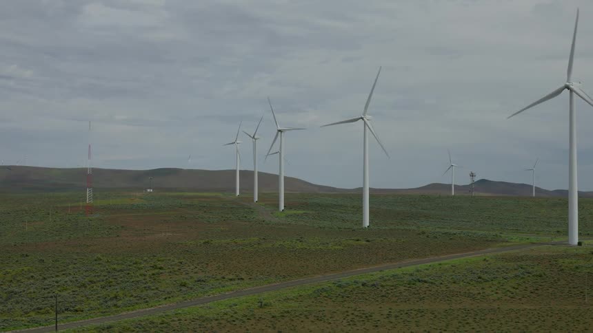 A long line of wind turbines spins slowly across a vast green and brown landscape under an overcast sky