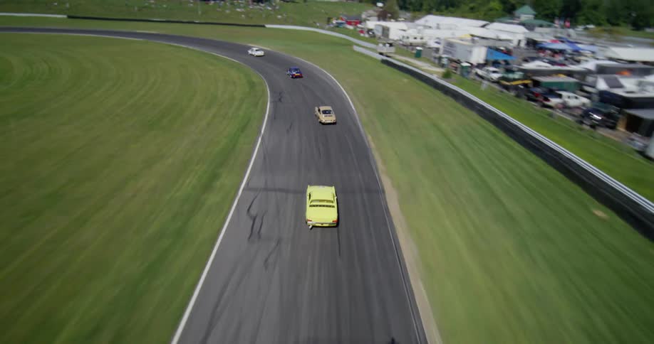 An aerial view follows vintage race cars as they speed around a curving racetrack on a sunny day