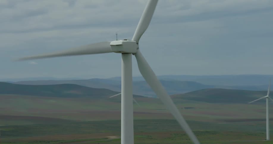 An aerial drone orbits a large, spinning wind turbine, revealing more turbines on a sprawling wind farm across rolling hills