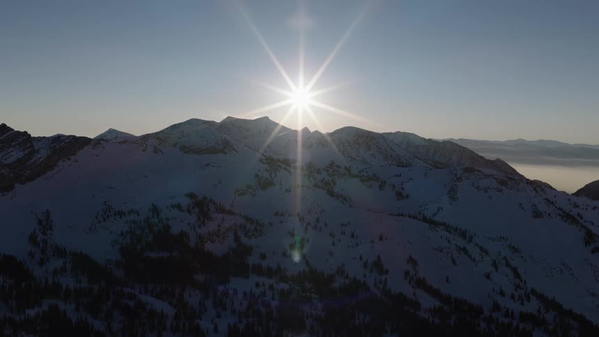 A bright sun shines like a star over a silhouetted, snow-covered mountain range