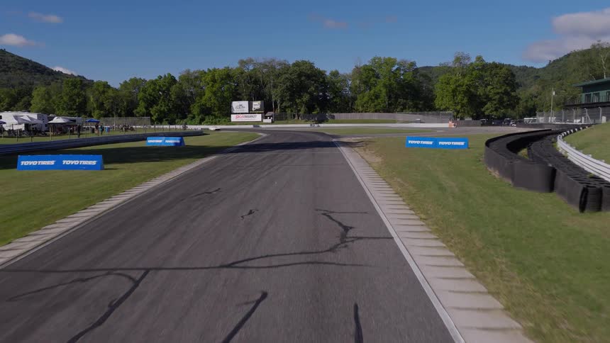 An aerial view follows vintage race cars as they speed under a bridge and around a scenic racetrack on a sunny day