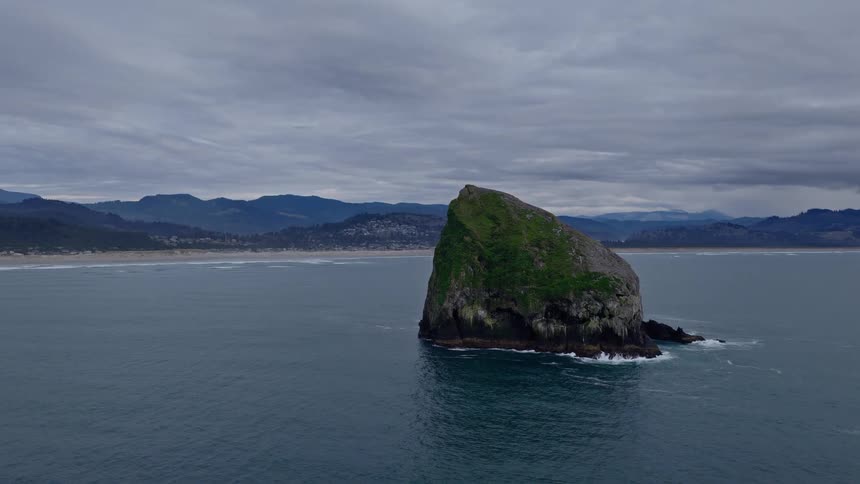A slow aerial pullback of a majestic, moss-covered sea stack isolated in the ocean off a rugged coastline under a cloudy sky