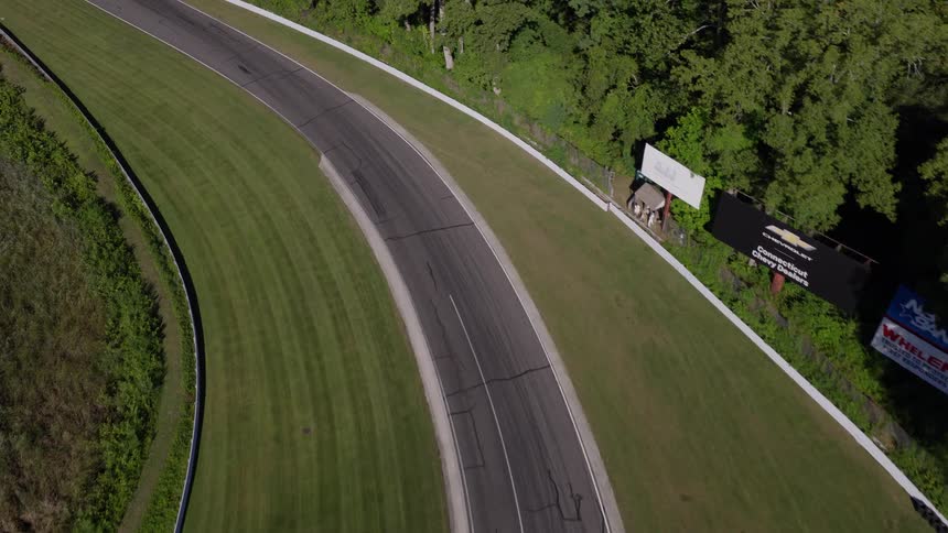 High-angle aerial shot follows several vintage race cars speeding down a curved track surrounded by green grass and trees