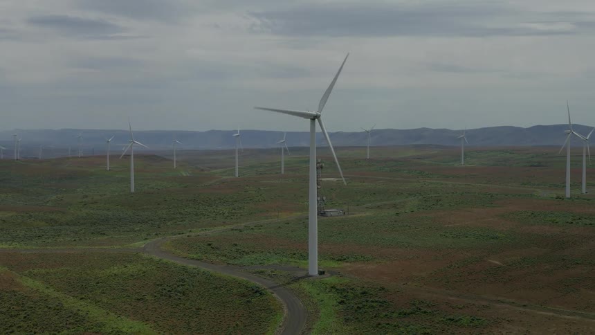 Slow aerial orbit of a massive wind turbine spinning on a sprawling wind farm set against a backdrop of rolling hills and a cloudy sky