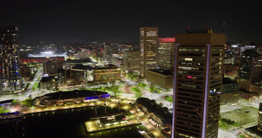 A cinematic aerial pan reveals the luminous Baltimore Inner Harbor and downtown skyline at night, with city lights reflecting on the water
