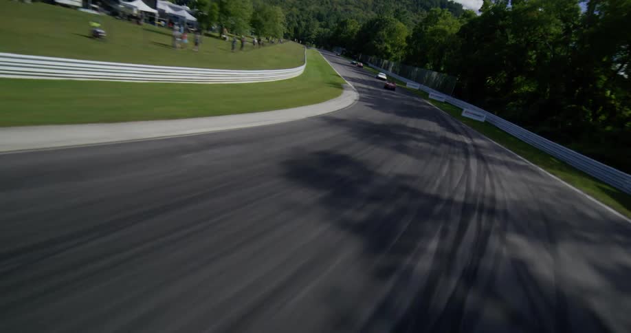 Dynamic aerial footage follows a pack of vintage sports cars as they race at high speed around a winding track in the countryside