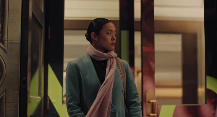 A stylish Asian woman in a teal coat and pink scarf waits patiently in a city doorway at night