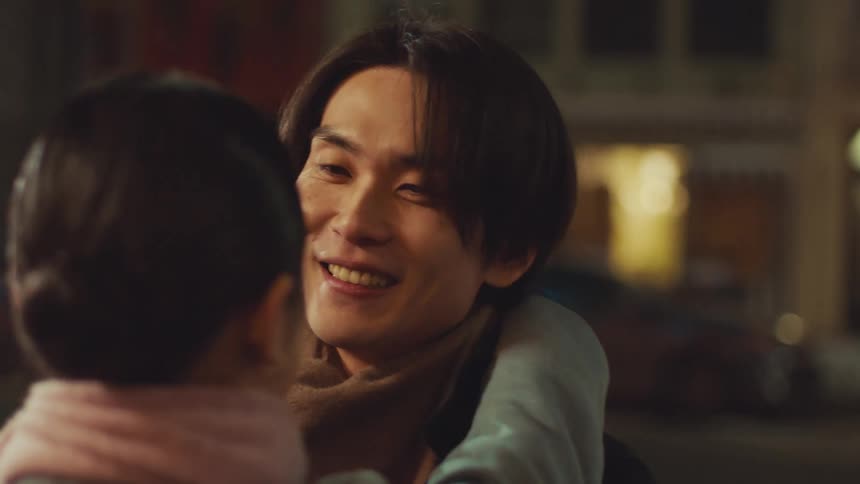A happy Asian man smiles as his girlfriend runs into his arms for a long, loving hug on a city street at night