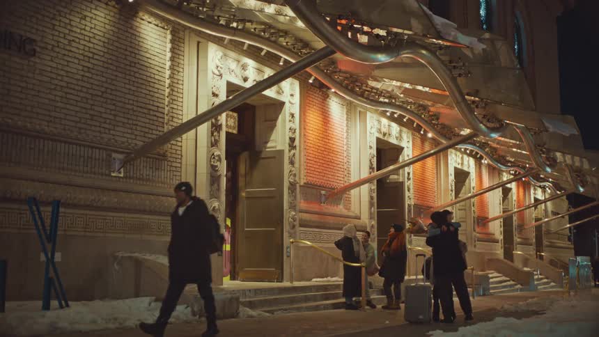 A diverse group of friends reunites with warm greetings and hugs outside an illuminated building on a snowy winter night