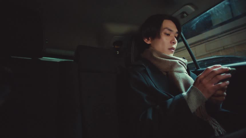 A young Asian man in a coat and scarf sits in the back of a moving car at night, intently using his smartphone