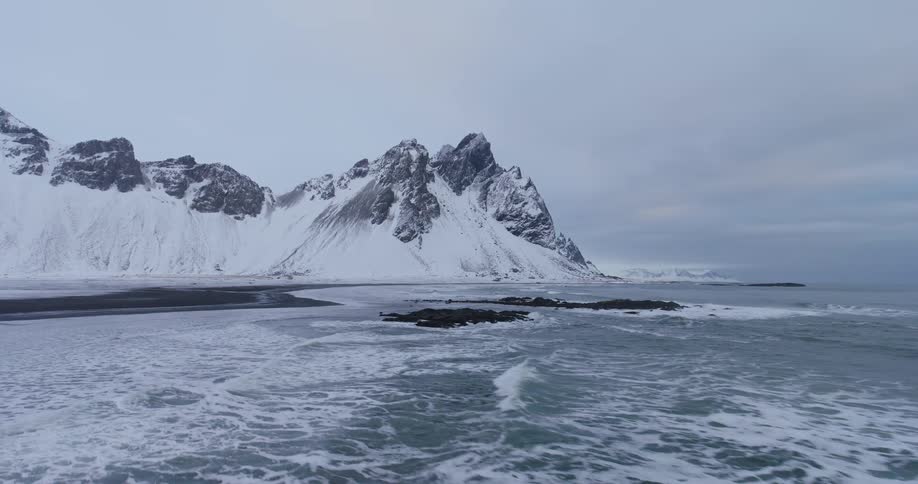 Icy ocean waves gently break along a snow-covered black sand beach at the base of a majestic mountain range in Iceland