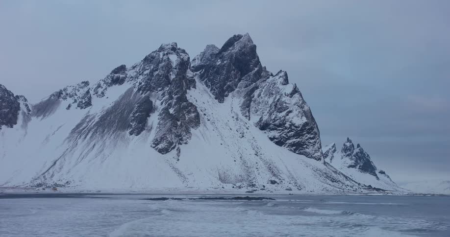 A majestic, snow-covered mountain range rises above an icy shoreline where gentle waves break