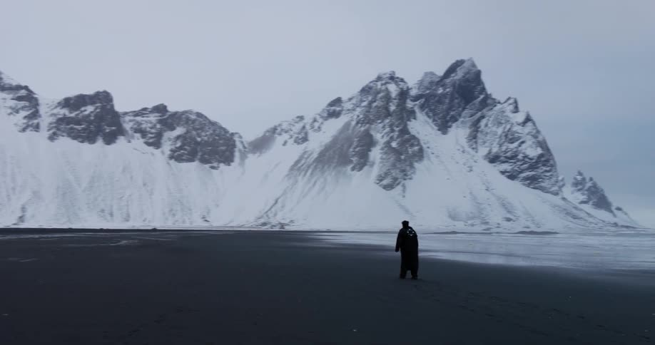 A solitary figure walks along a desolate black sand beach, dwarfed by the epic snow-covered mountains in the background on a cold, overcast day