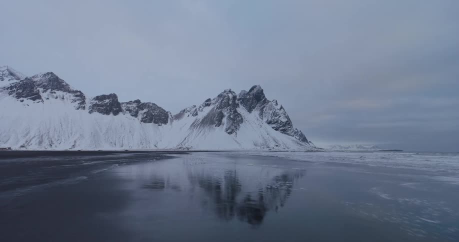 Gentle waves lap onto a black sand beach, creating a stunning reflection of a snow-covered mountain range