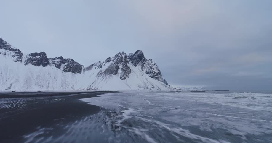 An aerial drone shot pulls back over the black sand beach and ocean, revealing the snow-covered Vestrahorn mountain in Iceland
