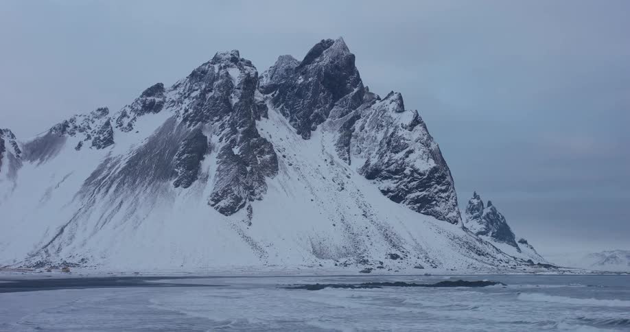 A majestic snow-covered mountain stands tall over a frozen, icy beach as ocean waves gently roll ashore