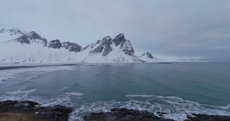 Epic aerial footage glides over the frigid ocean towards the iconic, snow-covered Vestrahorn mountain range on a winter day in Iceland