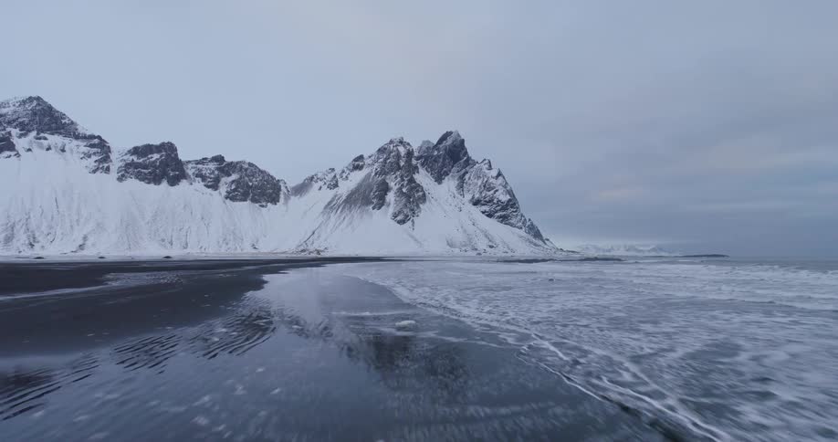 A serene aerial flight over an icy black sand beach reveals a perfect reflection of snow-capped mountains in the water