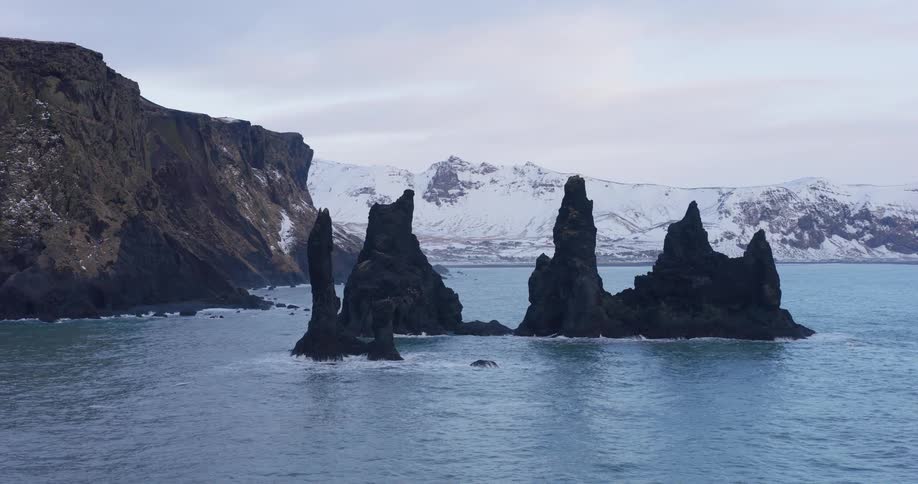 An epic aerial view reveals the majestic Reynisdrangar basalt sea stacks in the ocean along Iceland's stunning, snowy coastline during winter