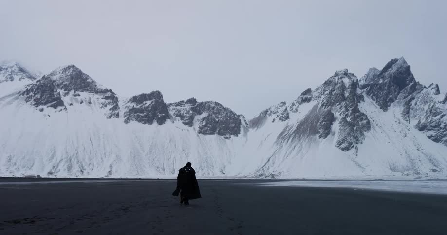 A lone figure in a dark cloak walks slowly across a vast, desolate landscape towards a majestic, snow-covered mountain range
