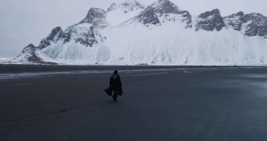 A lone figure in a dark cloak slowly walks across a vast black sand beach towards majestic snow-covered mountains in Iceland