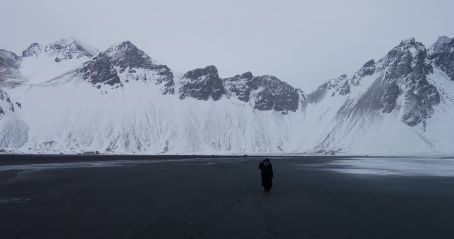 A lone figure in a long black coat walks across the vast black sand beach of Stokksnes with the epic, snow-covered Vestrahorn mountain in the background