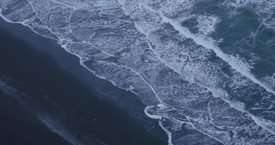 A dramatic top-down aerial view pulls back to reveal powerful ocean waves washing over a vast black sand beach