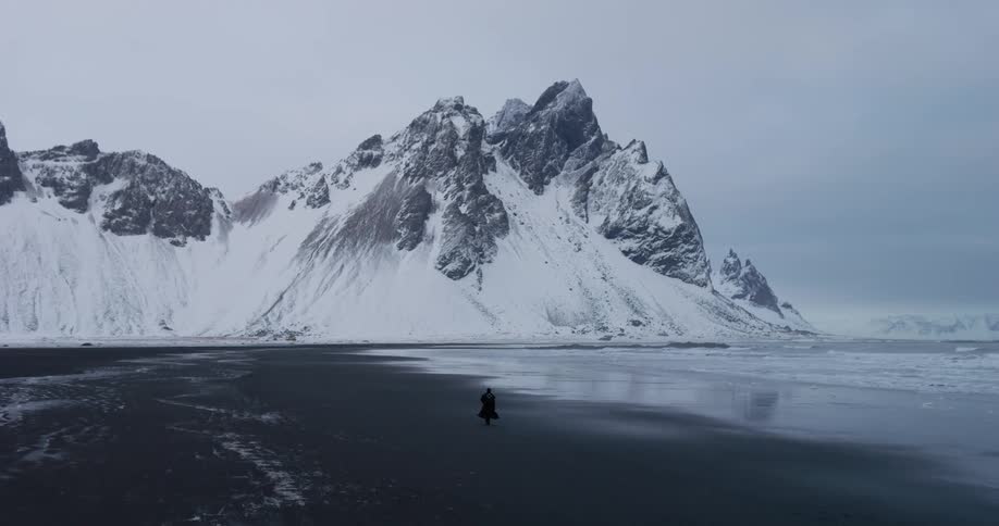 A solitary figure in a dark cloak walks across a vast black sand beach at the foot of Iceland's majestic, snow-covered Vestrahorn mountain