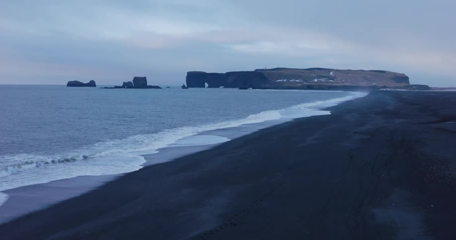 A dramatic aerial view of waves rolling onto a vast black sand beach along the iconic Icelandic coastline at twilight