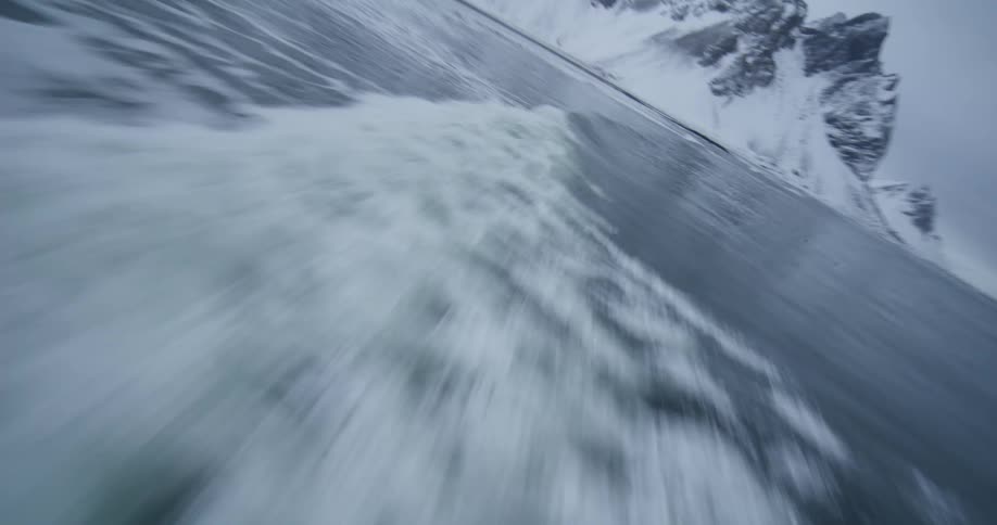 A stunning slow-motion FPV shot flies alongside a powerful ocean wave as it breaks towards a snowy mountain coastline