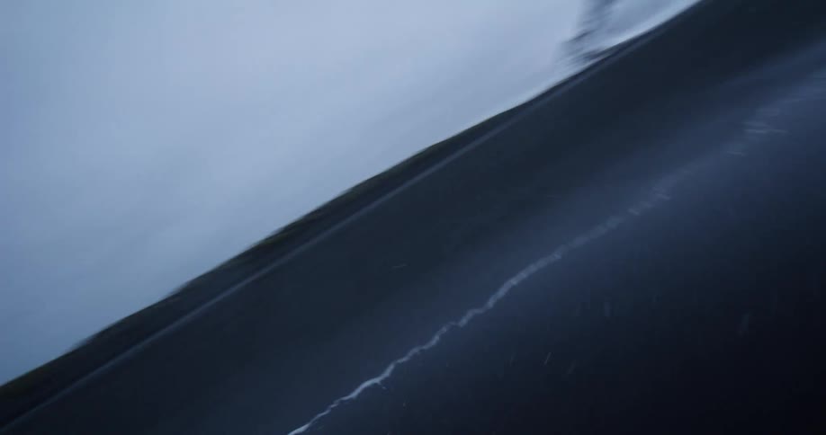 A surreal tilted shot follows a person running on a reflective black sand beach with dramatic, snow-covered mountains in the background