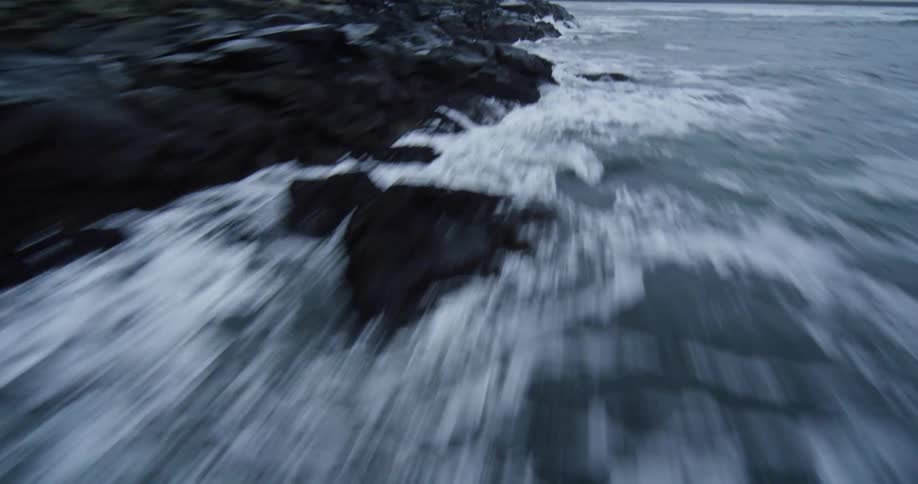 A dramatic, fast-moving aerial shot skims low over the ocean waves as they crash against a dark, rocky shoreline