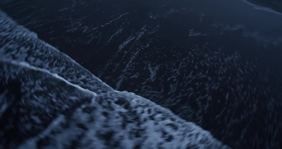 Dynamic low-angle tracking shot of ocean waves washing over a moody black sand beach with a snowy landscape in the background