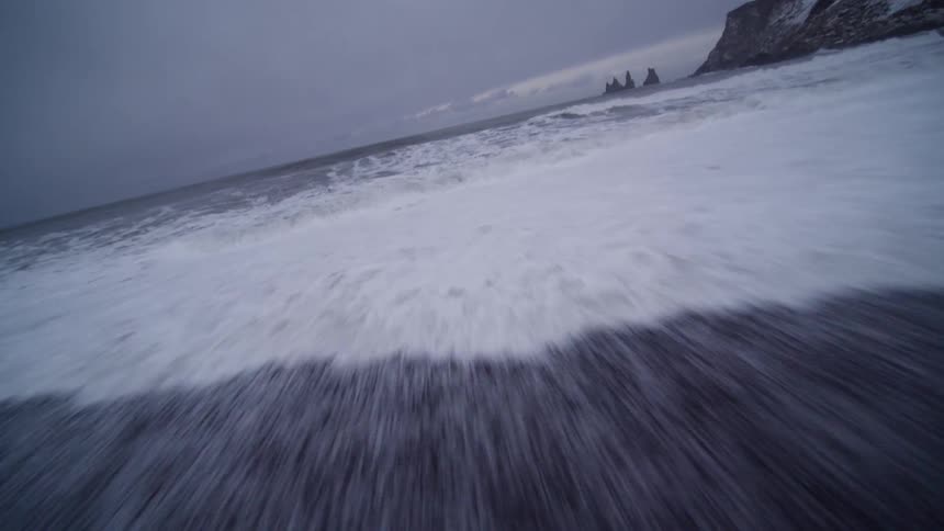 A thrilling low-angle point-of-view shot skims across a snowy black sand beach and plunges into the turbulent, crashing ocean waves