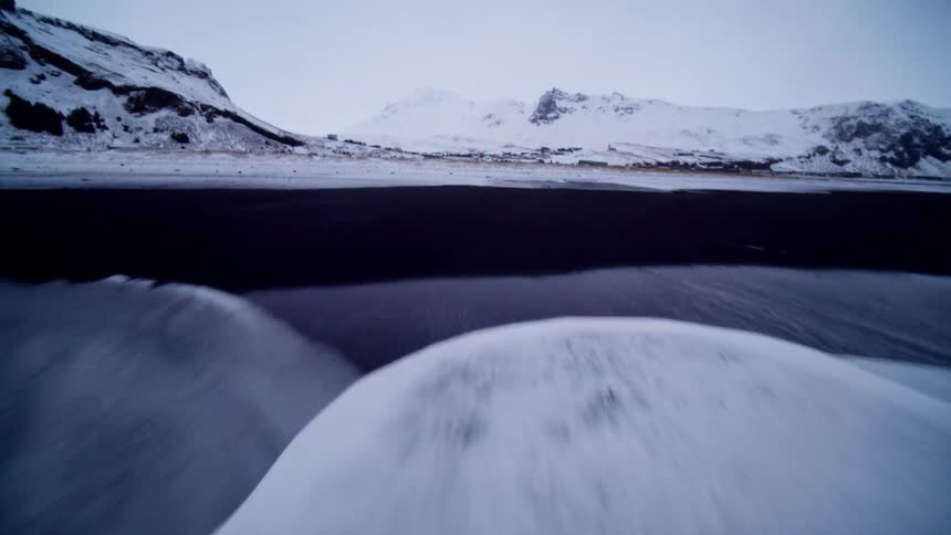 An immersive aerial shot flies backward from the frothy ocean waves over a snow-covered black sand beach in Iceland