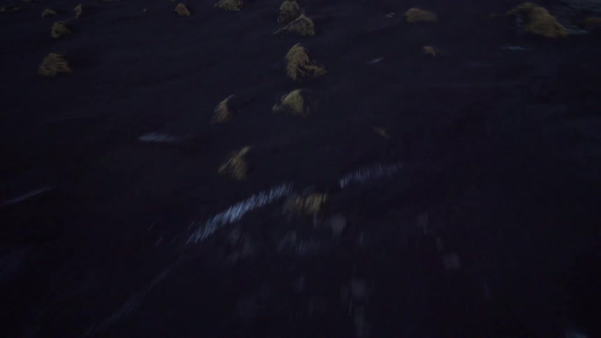 A fast-paced drone shot soars over a desolate black sand beach dotted with patches of snow and grass mounds at dusk