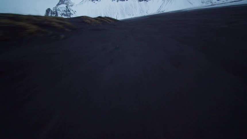 Skimming across a desolate black sand beach toward snow-covered mountains in a fast-paced aerial shot