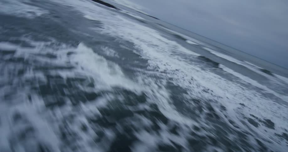 An immersive FPV shot races along the crest of a dark ocean wave as it breaks towards a snowy arctic coastline