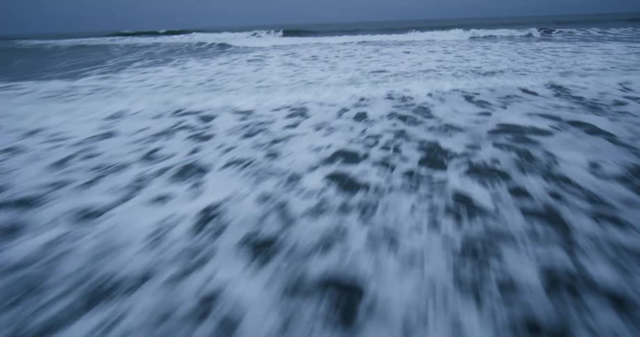 A fast-paced, low-angle FPV-style shot skims across the surface of dark, choppy ocean waves under an overcast sky