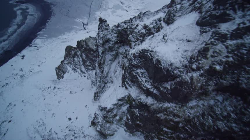 A dramatic top-down aerial flight along a jagged, snow-covered mountain ridge reveals the cold ocean waves below