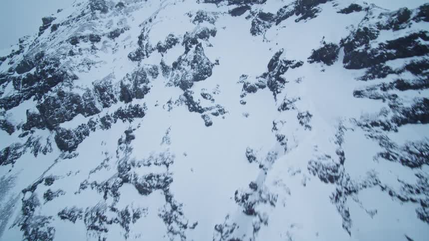 A slow aerial drone shot ascends the steep, rugged face of a snow-covered mountain on an overcast, foggy day
