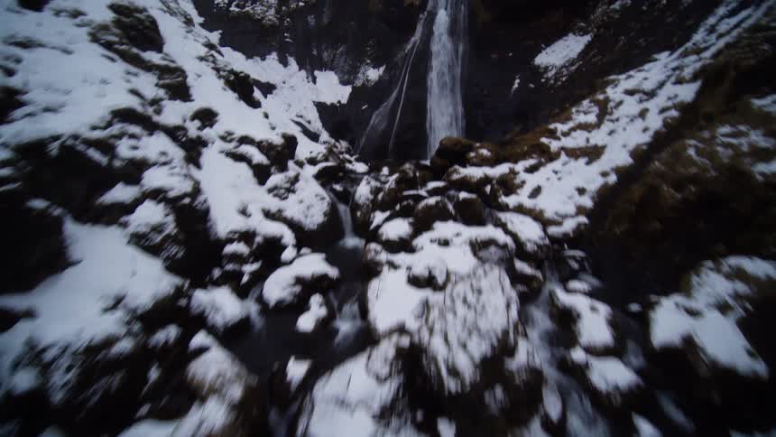 Dynamic FPV drone footage soars up a snow-covered river gorge, revealing a series of cascading waterfalls in a rocky, wintery canyon